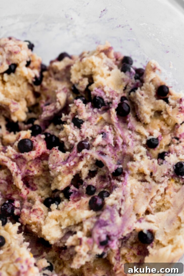 Frozen wild blueberries gently folded into the yellow cookie dough in a glass bowl, showing their vibrant color.