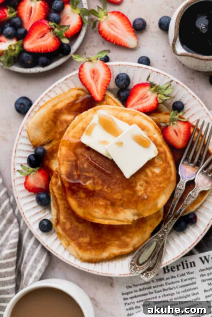 Heavenly Sweet Cream Pancakes 2 Close up of a stack of golden brown sweet cream pancakes on a white plate, garnished with fresh strawberries and blueberries.