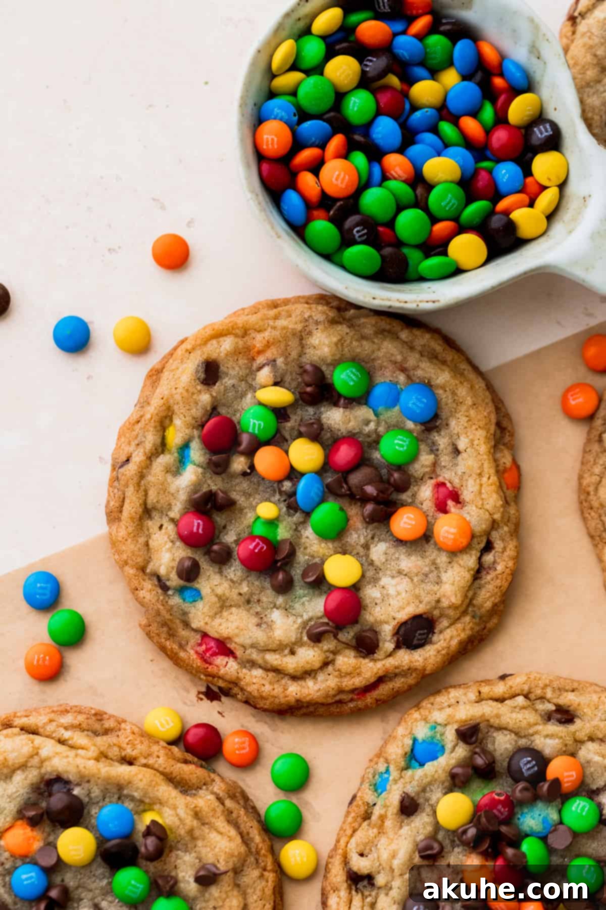 One freshly baked mini M&M cookie rests on parchment paper next to a bowl brimming with colorful mini M&Ms, ready for baking.