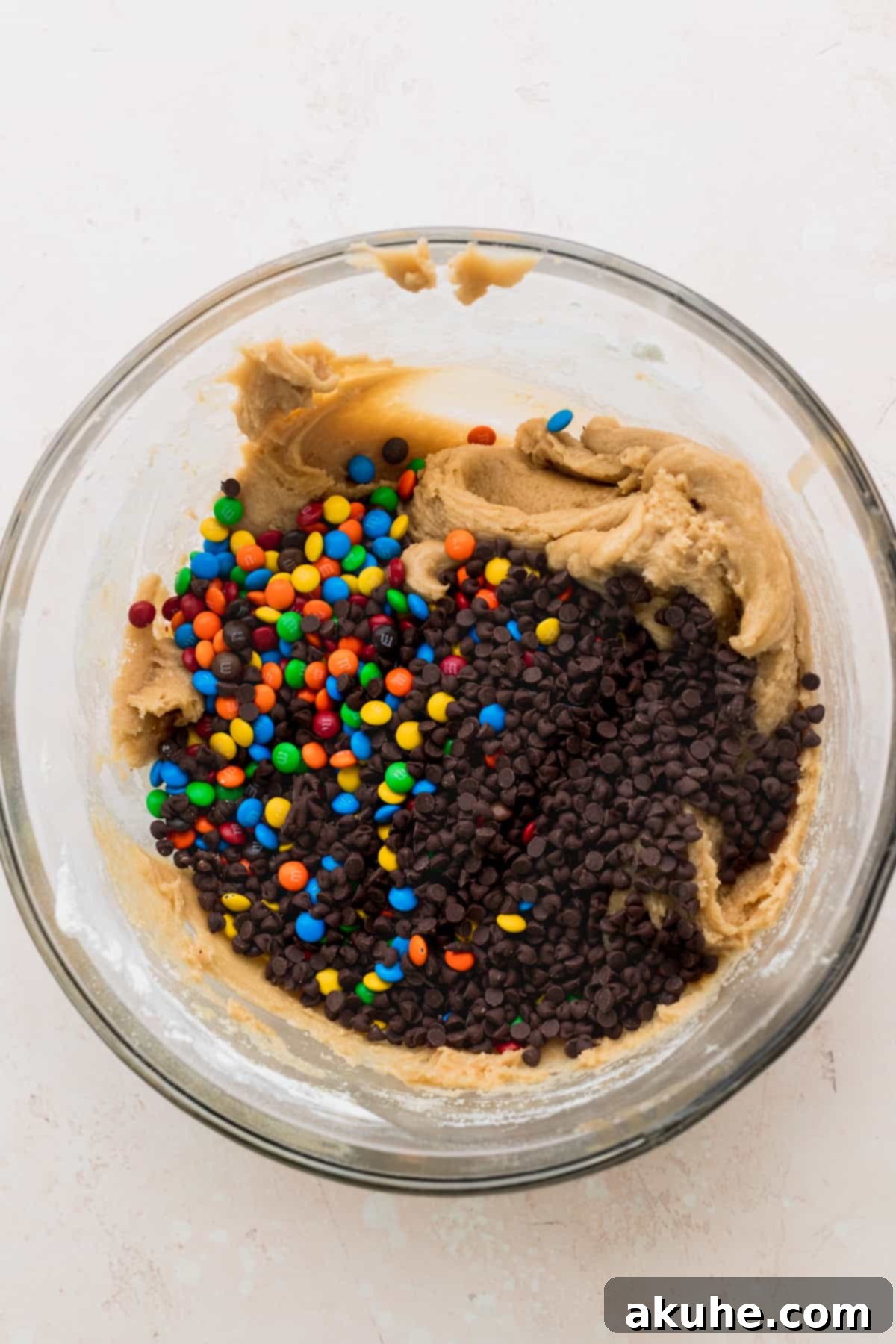 Cookie dough in a glass bowl, generously topped with an assortment of colorful mini M&Ms, ready to be folded in.