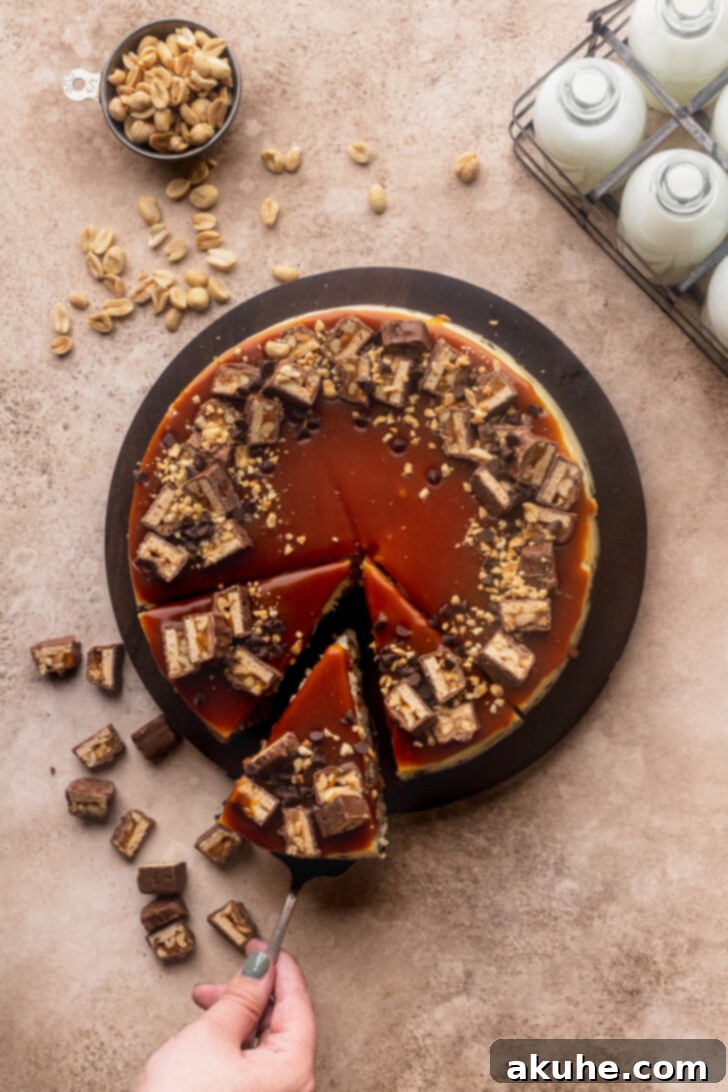 Snickers Bar Cheesecake Perfection 7 A fully decorated Snickers cheesecake resting on a wooden cutting board, topped with extra Snickers, peanuts, and mini chocolate chips.