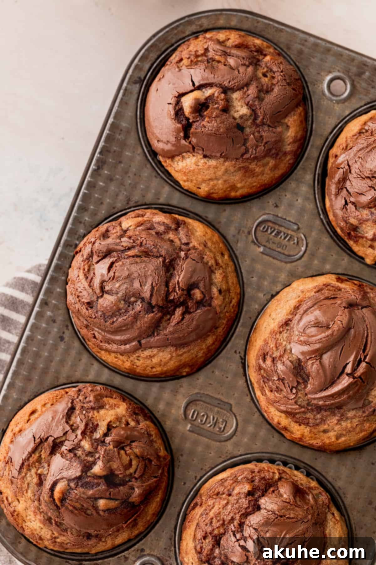 Nutella Swirl Banana Muffins 2 Freshly baked Nutella banana muffins cooling in a muffin pan, showcasing their golden tops and visible Nutella swirls.