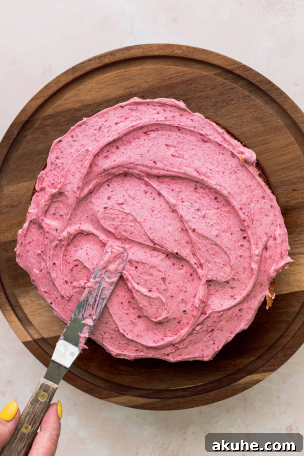 An offset spatula expertly spreading a thick layer of creamy raspberry frosting onto a vanilla cake layer, forming the base for the filling.