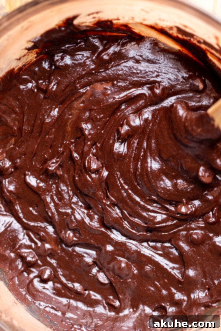 Thick brownie batter in a mixing bowl with chocolate chips being folded in using a rubber spatula.