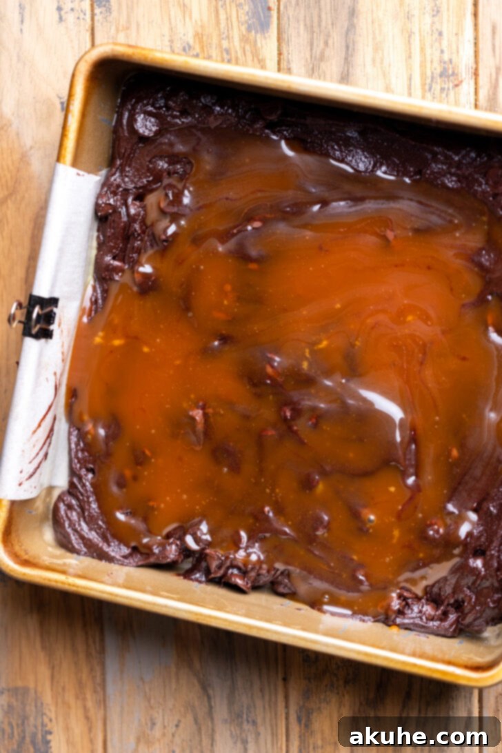 Gooey salted caramel being spread over a layer of brownie batter in a baking pan with an offset spatula.