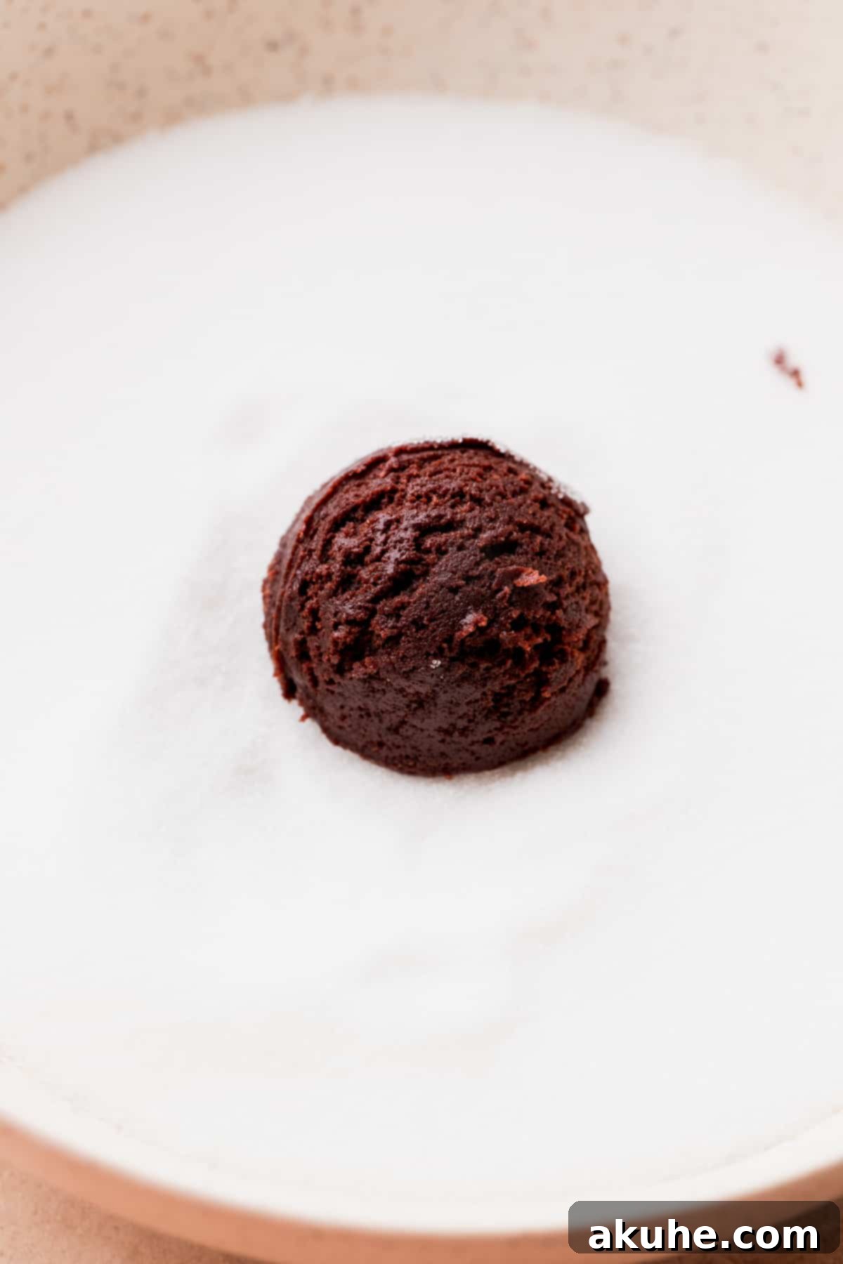 Warm Cocoa Blossom Cookies 6 A perfectly round chocolate cookie dough ball being rolled in a bowl of white granulated sugar, creating an even coating.