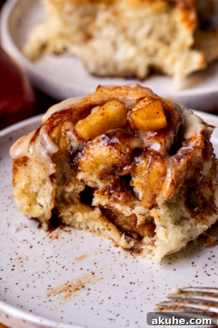 Homemade Apple Cinnamon Rolls 2 Bite missing from an irresistible apple cinnamon roll on a plate, showcasing its gooey center and creamy icing.