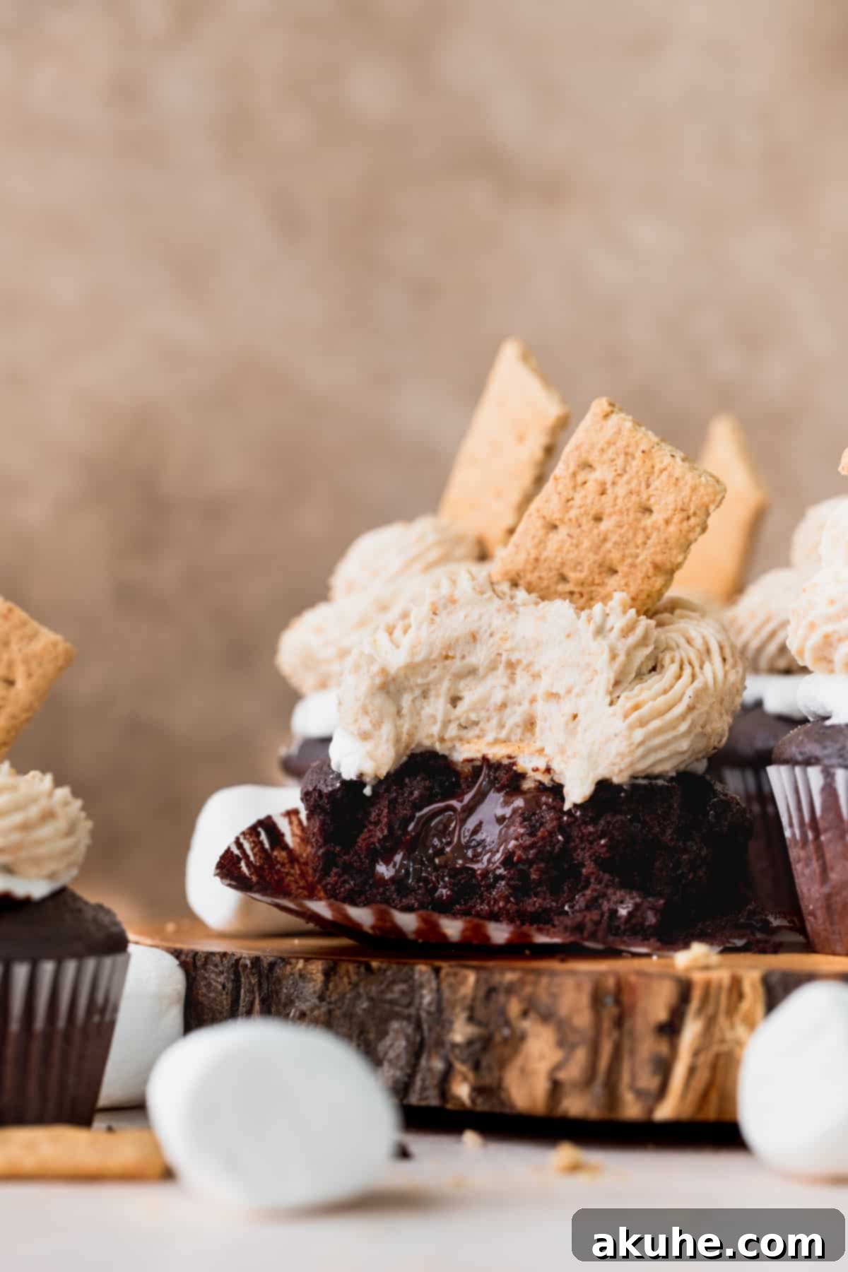 A s'mores cupcake with a bite taken out, revealing the chocolate ganache filling and fluffy toasted marshmallow topping, resting on a wooden board amidst other whole cupcakes.