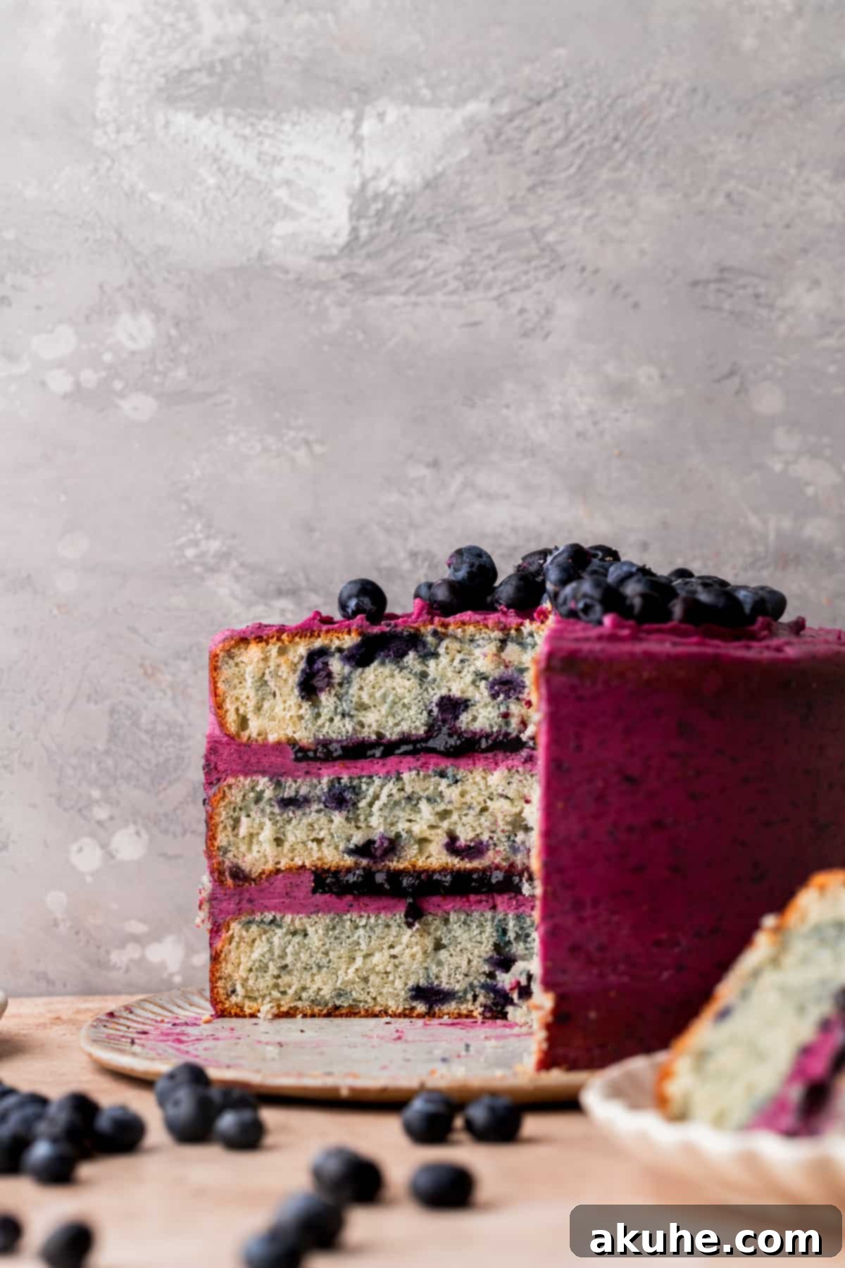 Bursting Berry Bliss 2 A slice of triple blueberry cake with visible layers of vanilla cake, blueberry sauce, and blueberry frosting, elegantly presented on a platter.