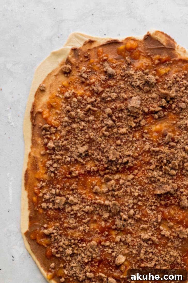 Peach Cobbler Cinnamon Rolls Reimagined 7 Crumbly cinnamon topping sprinkled evenly over the peach jam and cinnamon filling on the dough, adding texture.