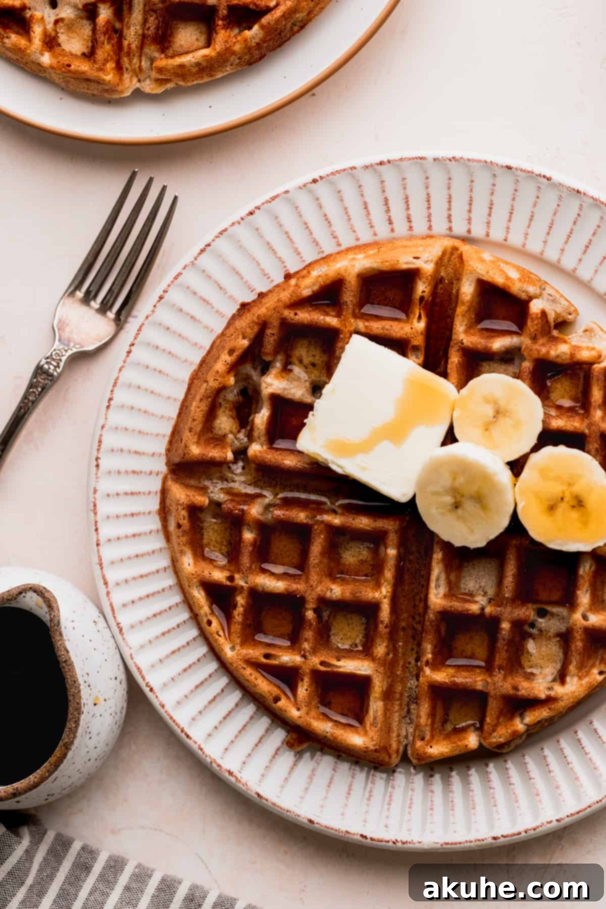 Golden-brown banana waffles on a white plate, adorned with a pat of melting butter, a drizzle of maple syrup, and slices of fresh banana.