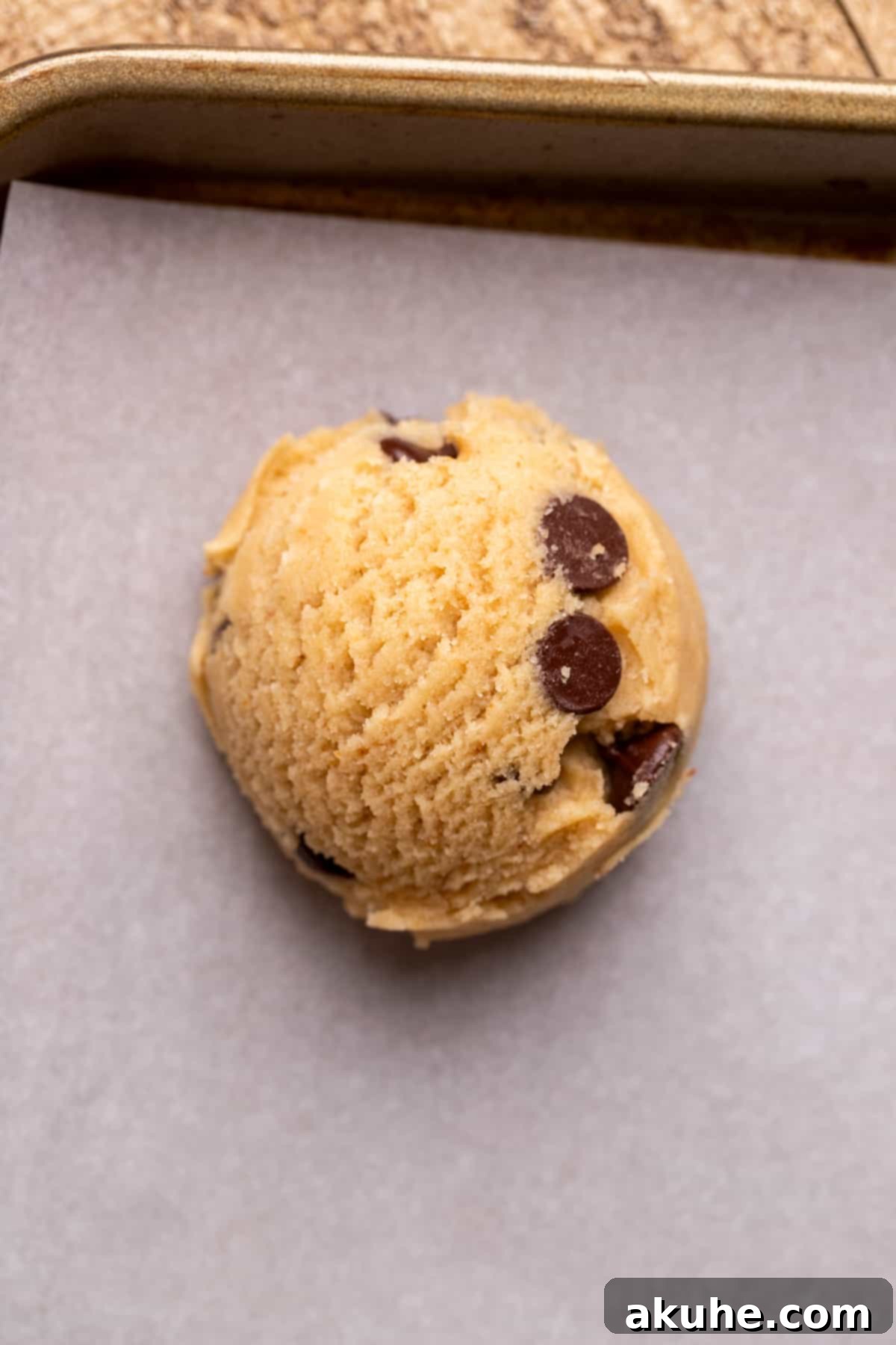 A single, perfectly scooped ball of chocolate chip cookie dough resting on parchment paper on a baking sheet, ready for the oven.