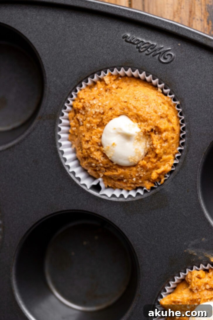 Cream cheese filling being piped into the center of pumpkin muffin batter in a muffin liner, creating a swirl.