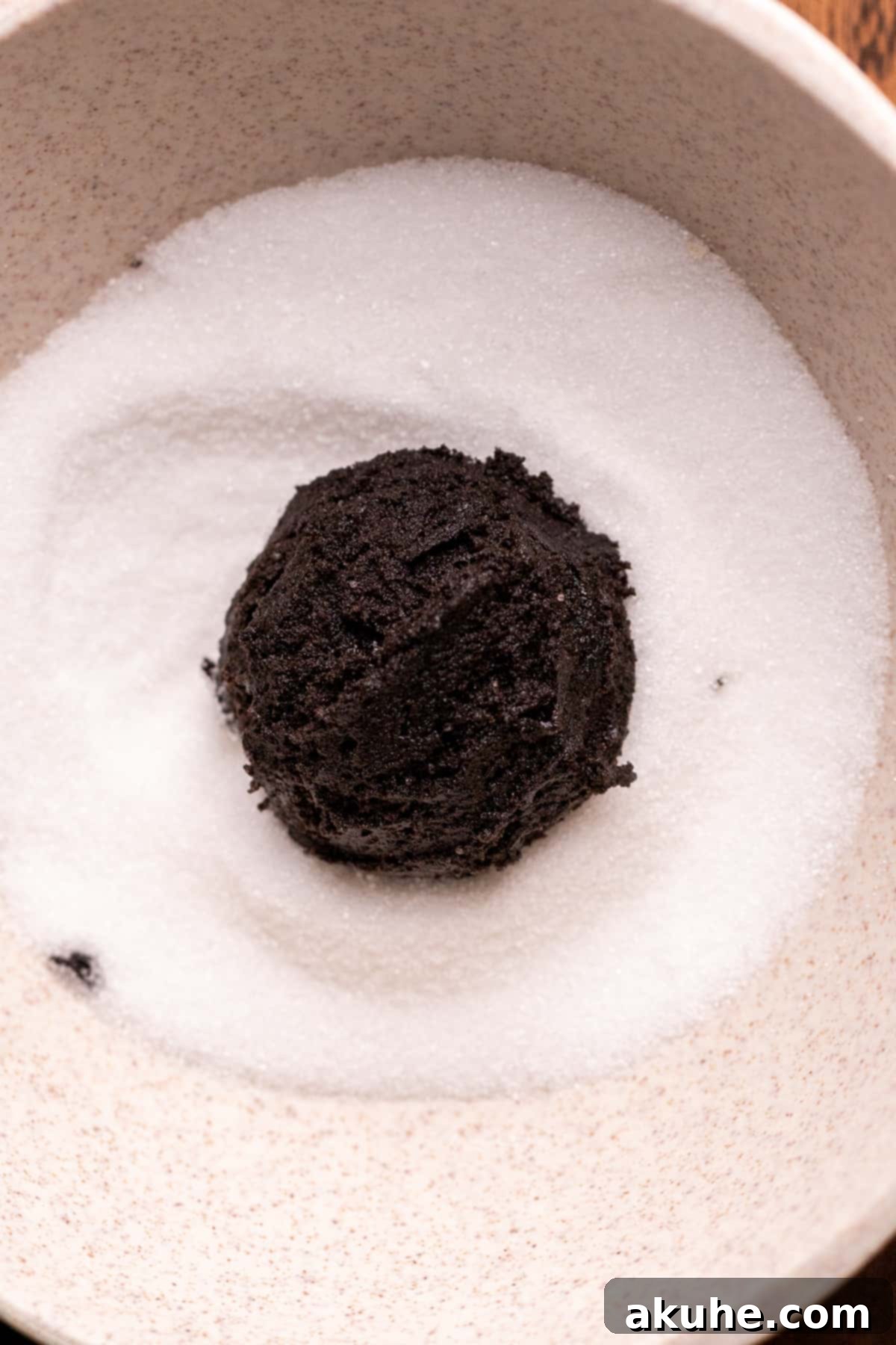 A ball of black cocoa cookie dough being rolled in a bowl of white granulated sugar.