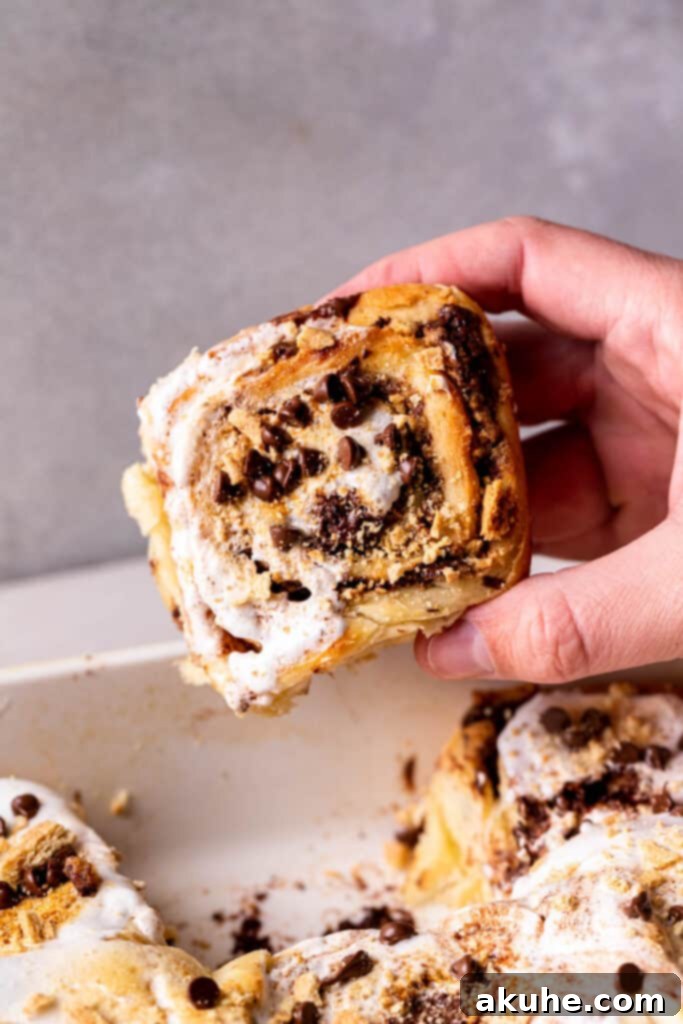 A hand gently reaches for a warm S'mores cinnamon roll, glistening with toasted marshmallow and melted chocolate, from a baking pan.