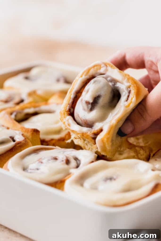 A hand gently lifts a warm, generously frosted homemade cinnamon roll from a baking pan, with steam softly rising, highlighting its gooey center.