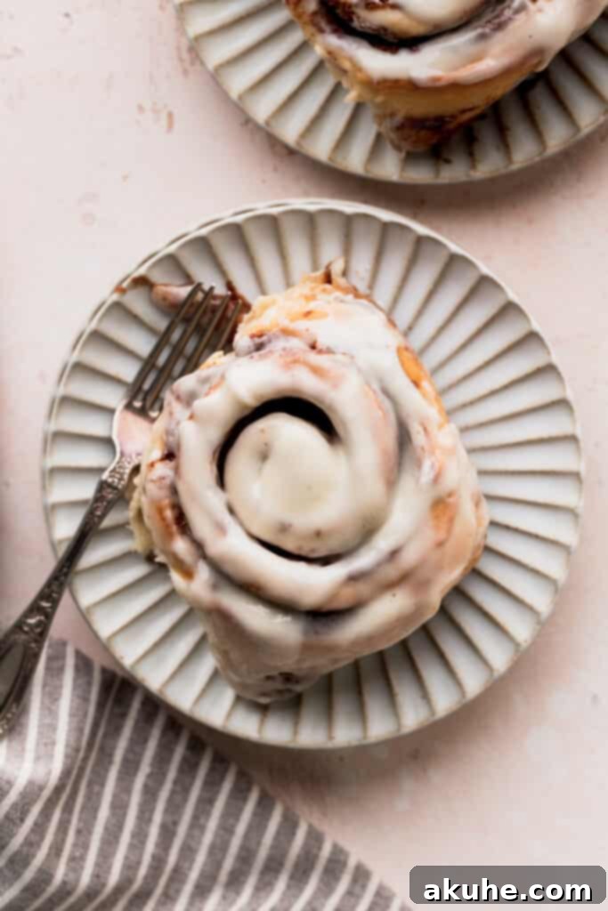 A single, generously frosted giant cinnamon roll rests on a pristine white plate, with a fork poised, ready for an indulgent experience.