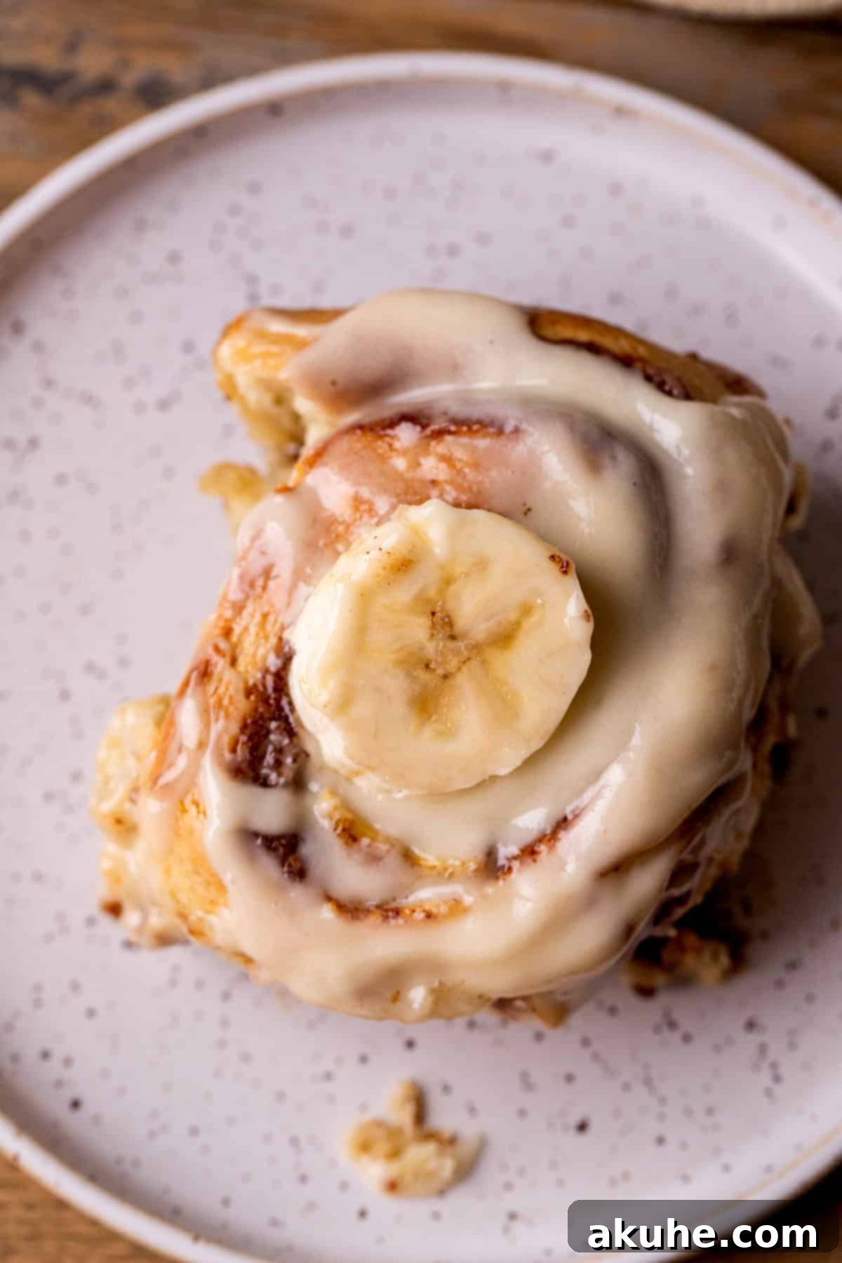Banana Swirl Buns 9 A single Banana Cinnamon Roll with a generous layer of cream cheese icing on a white plate, viewed from above.