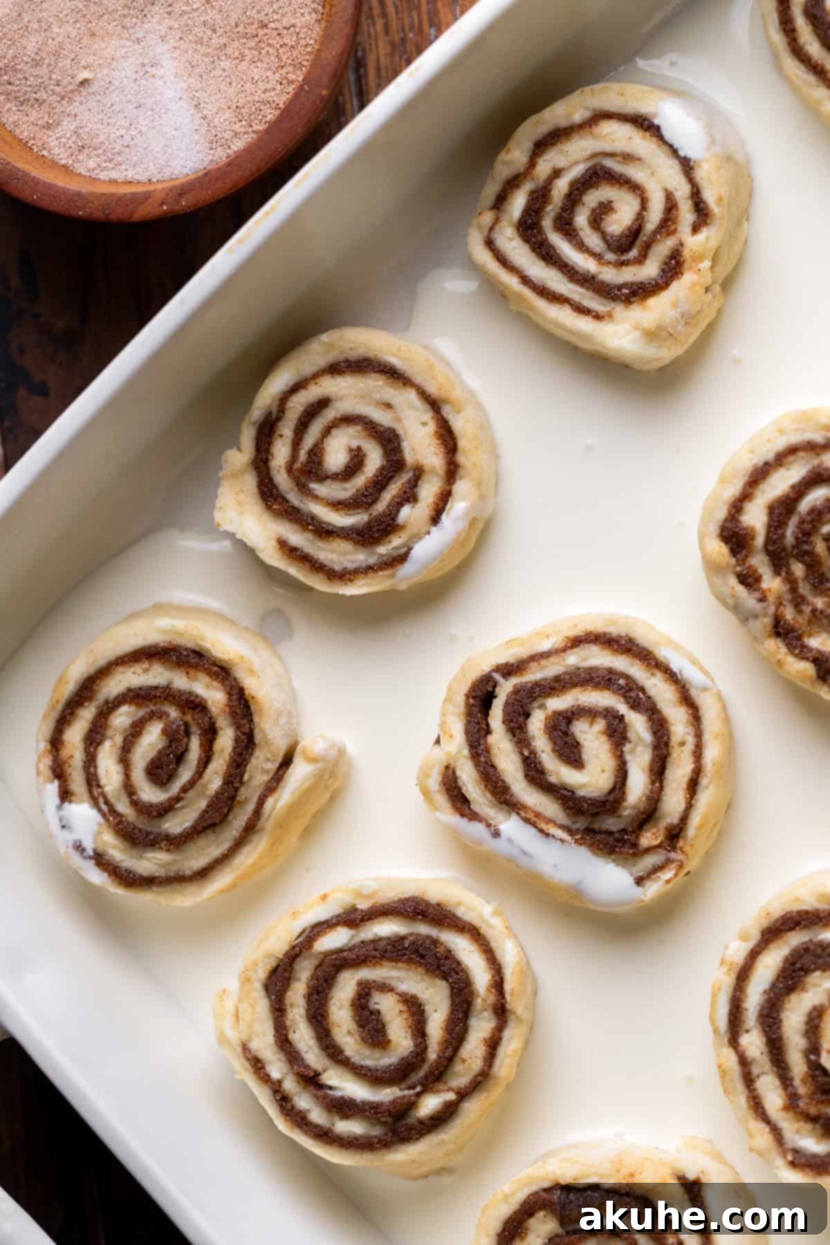 Heavy cream being poured around the unbaked cinnamon rolls in a glass baking pan.