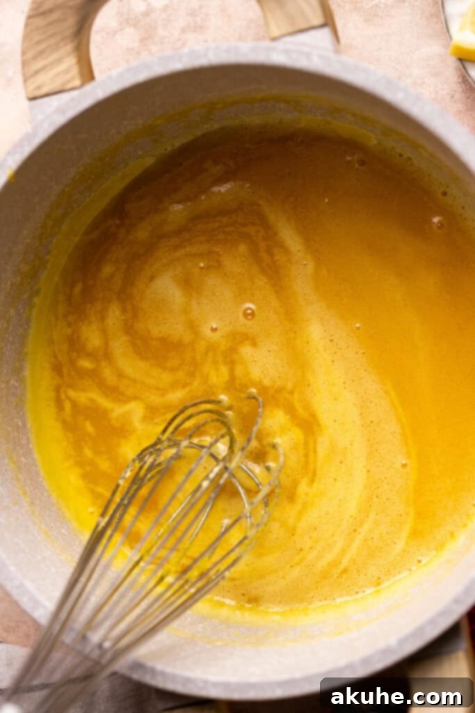 The lemon curd mixture in a pot has thickened significantly, coating the whisk and indicating it's nearly at the perfect stage before the final steps.