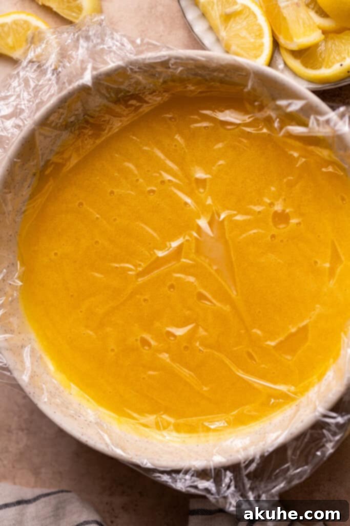 A bowl of freshly prepared lemon curd is shown covered tightly with clear plastic wrap, ensuring the wrap touches the surface to prevent any skin from forming during the cooling process.