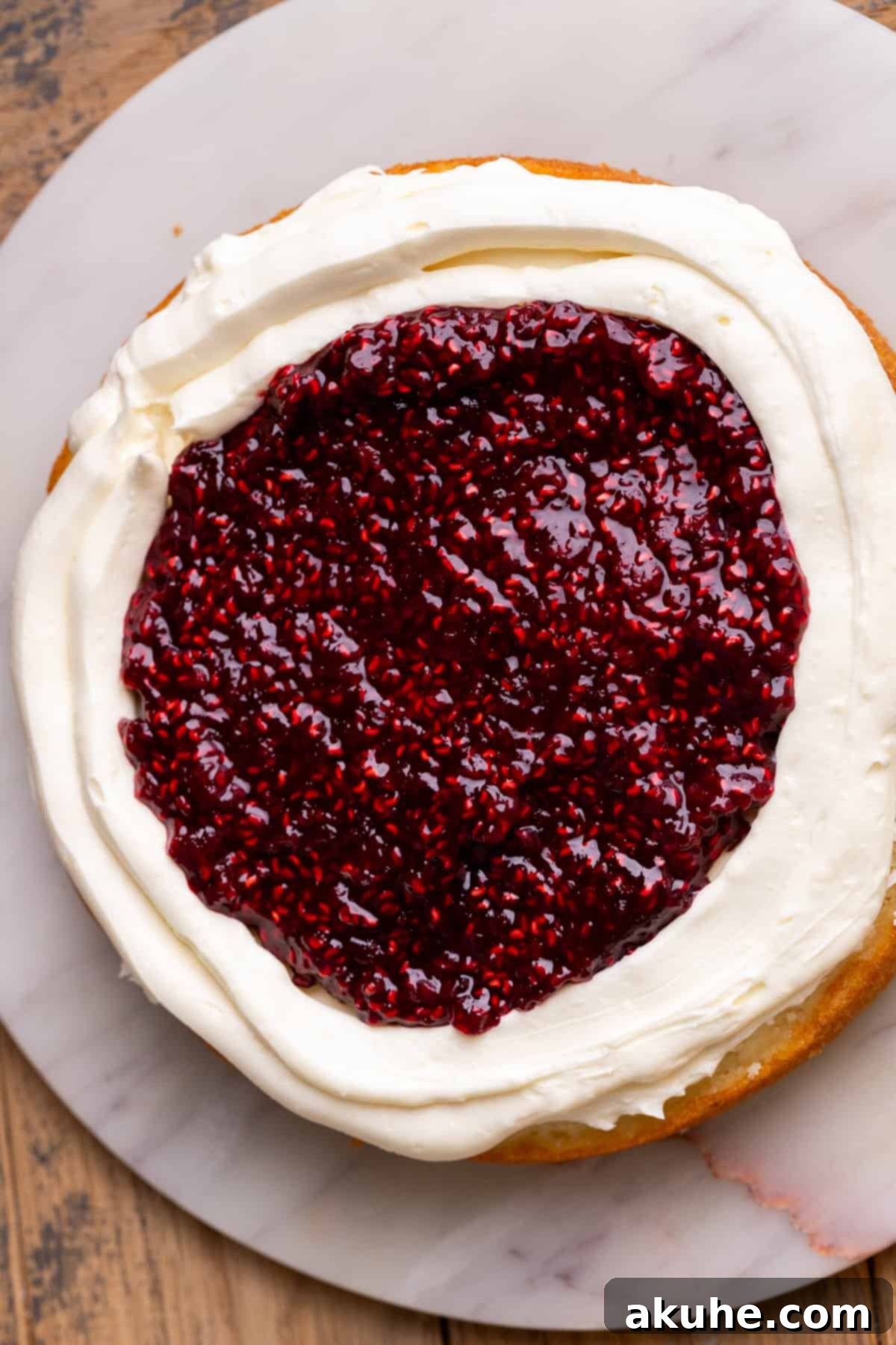 A cake layer topped with white chocolate frosting and a piped border, filled with raspberry jam.