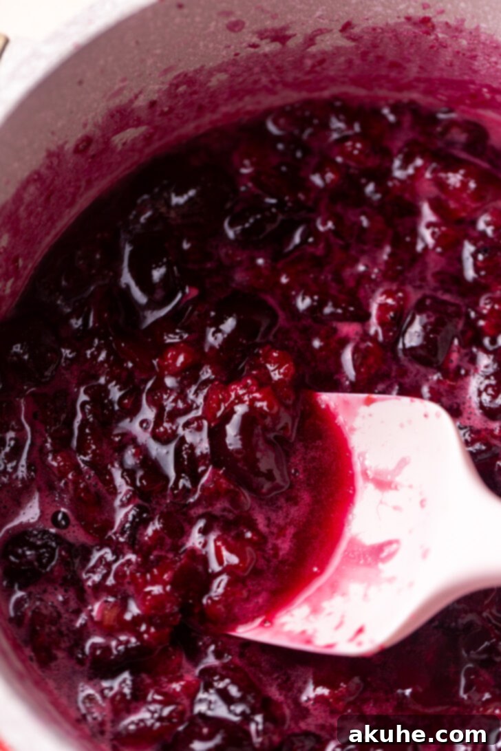 Homemade Cherry Pie Filling 5 Cherries simmering in a pot on the stove, with a potato masher partially submerged, indicating the process of mashing to achieve the desired texture.