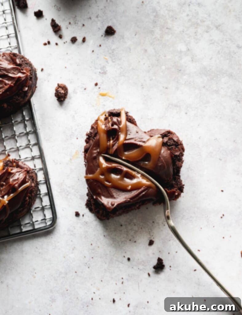 A single heart-shaped brownie, topped with ganache and caramel, with a fork gently inserted, showing its rich, soft interior.