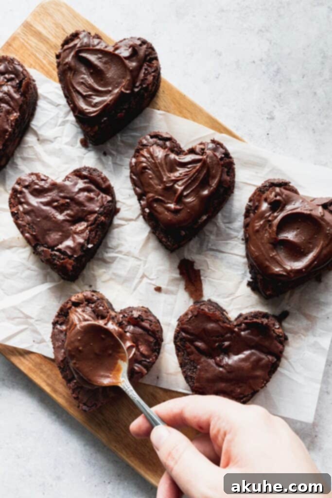 A spoon spreading rich, dark chocolate ganache smoothly over the top of a heart-shaped brownie.