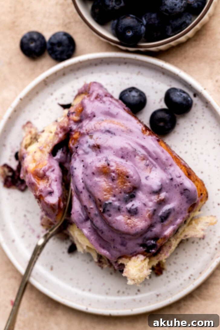 Close-up top view of a single blueberry cinnamon roll on a white plate, showing the creamy icing and blueberry swirls.