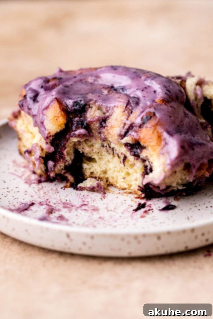 Side view of a fluffy blueberry cinnamon roll with a bite taken, showcasing the layers of dough, cinnamon, and blueberry jam, topped with cream cheese icing.
