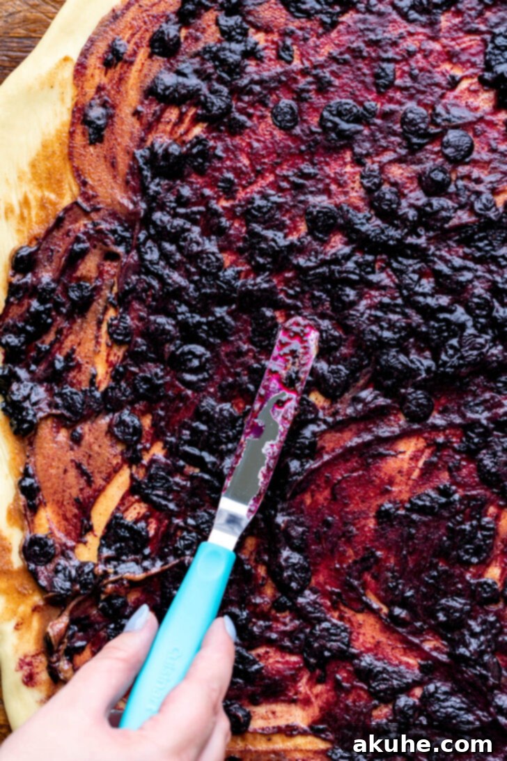 Blueberry jam being spread over the cinnamon filling, reaching to the edges of the dough.