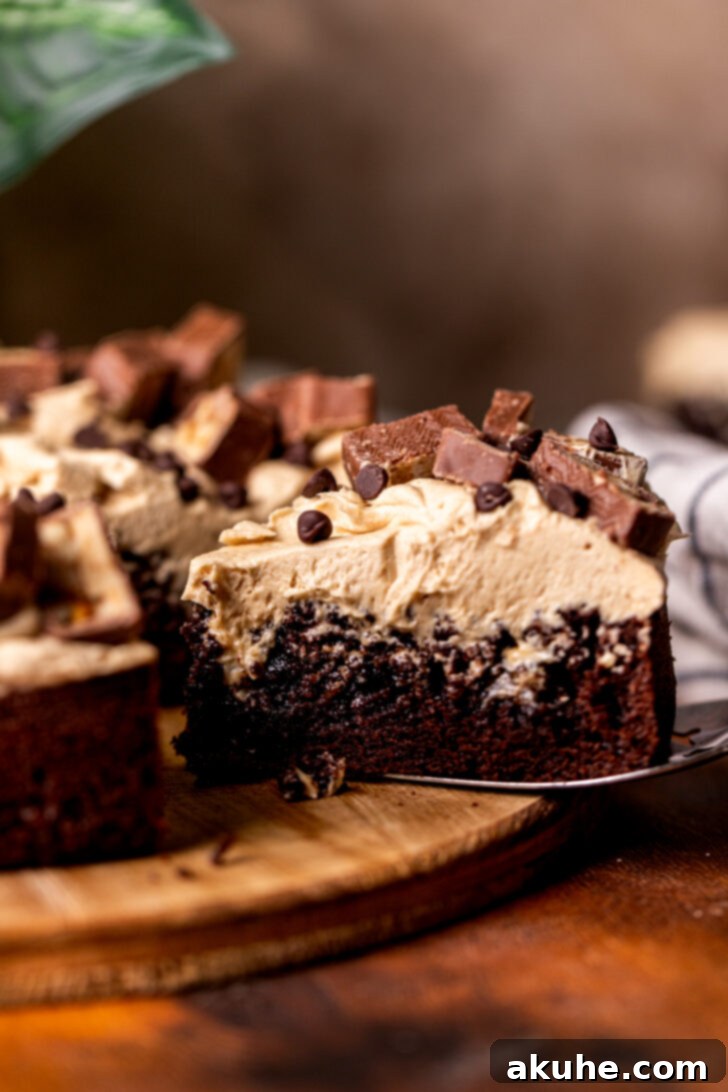 A generous slice of Snickers cake being removed from the main cake, showcasing its layers of chocolate cake, caramel filling, peanut butter frosting, and Snickers topping.