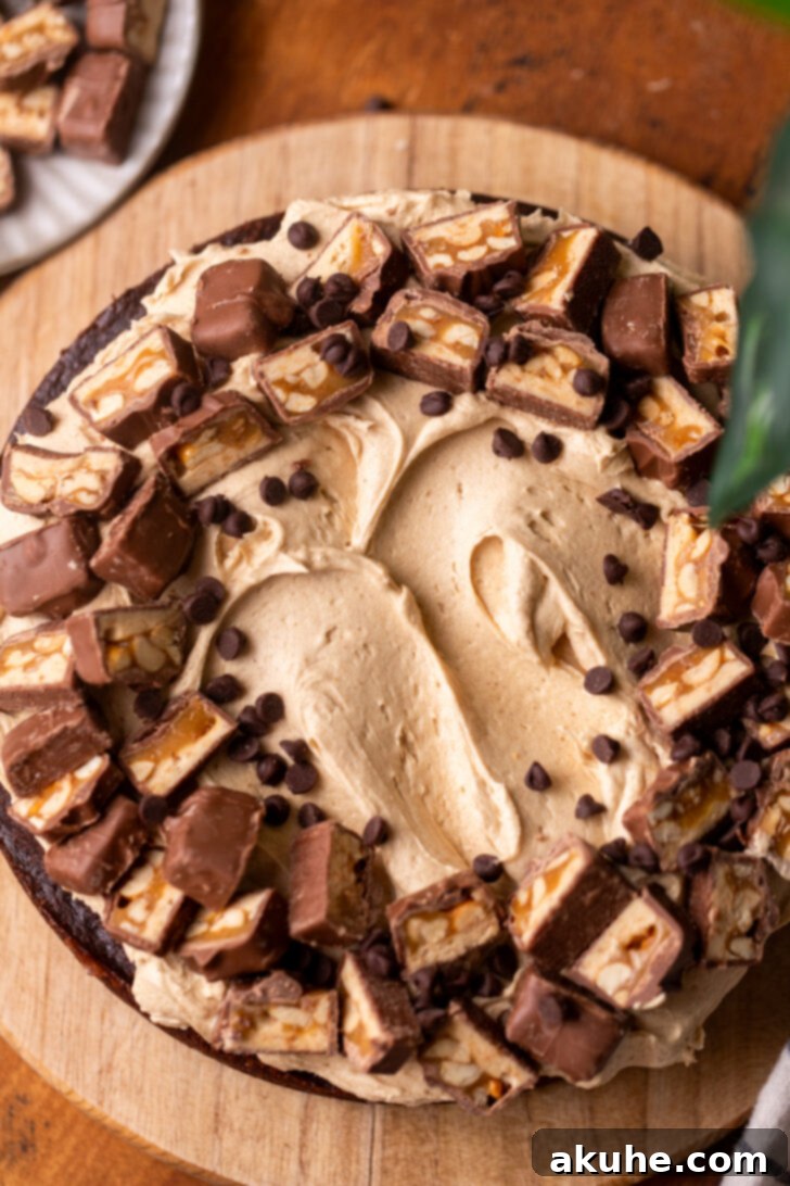 The fully decorated Snickers cake on a wooden serving board, topped with peanut butter frosting and chopped Snickers candy.