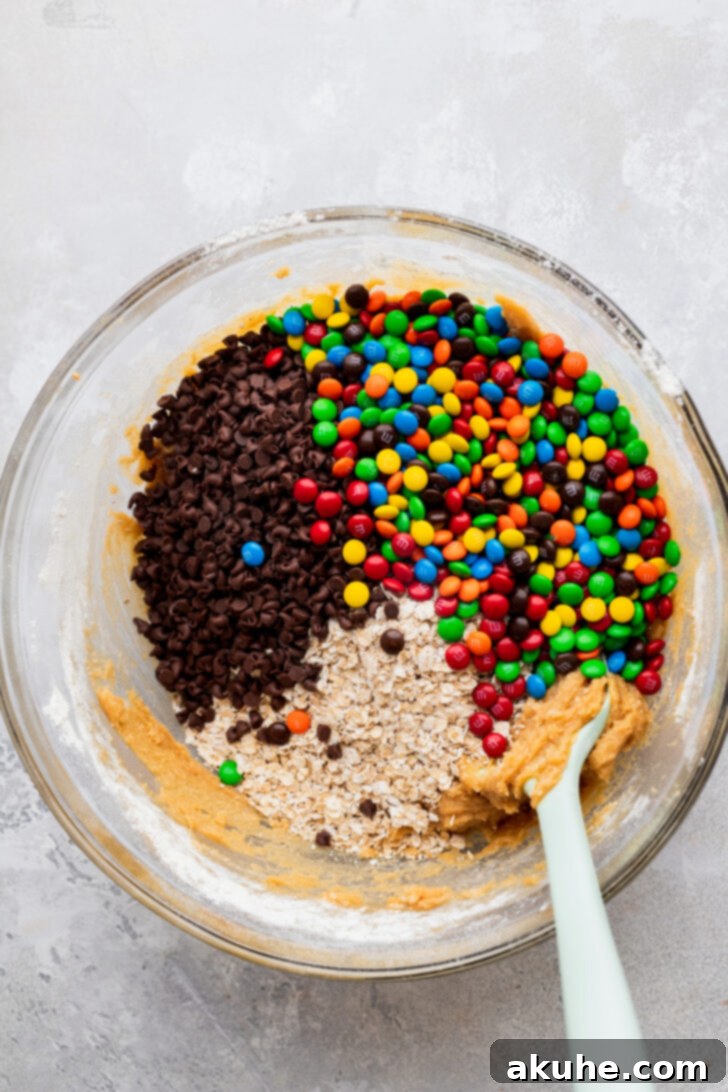Kitchen Sink Cookies 5 Cookie dough with dry ingredients added, then oats, mini chocolate chips, and mini M&Ms being gently folded in with a rubber spatula.