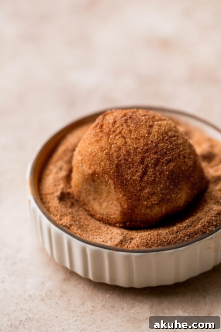 A ball of cookie dough being rolled in a bowl of cinnamon sugar, ensuring a generous coating.