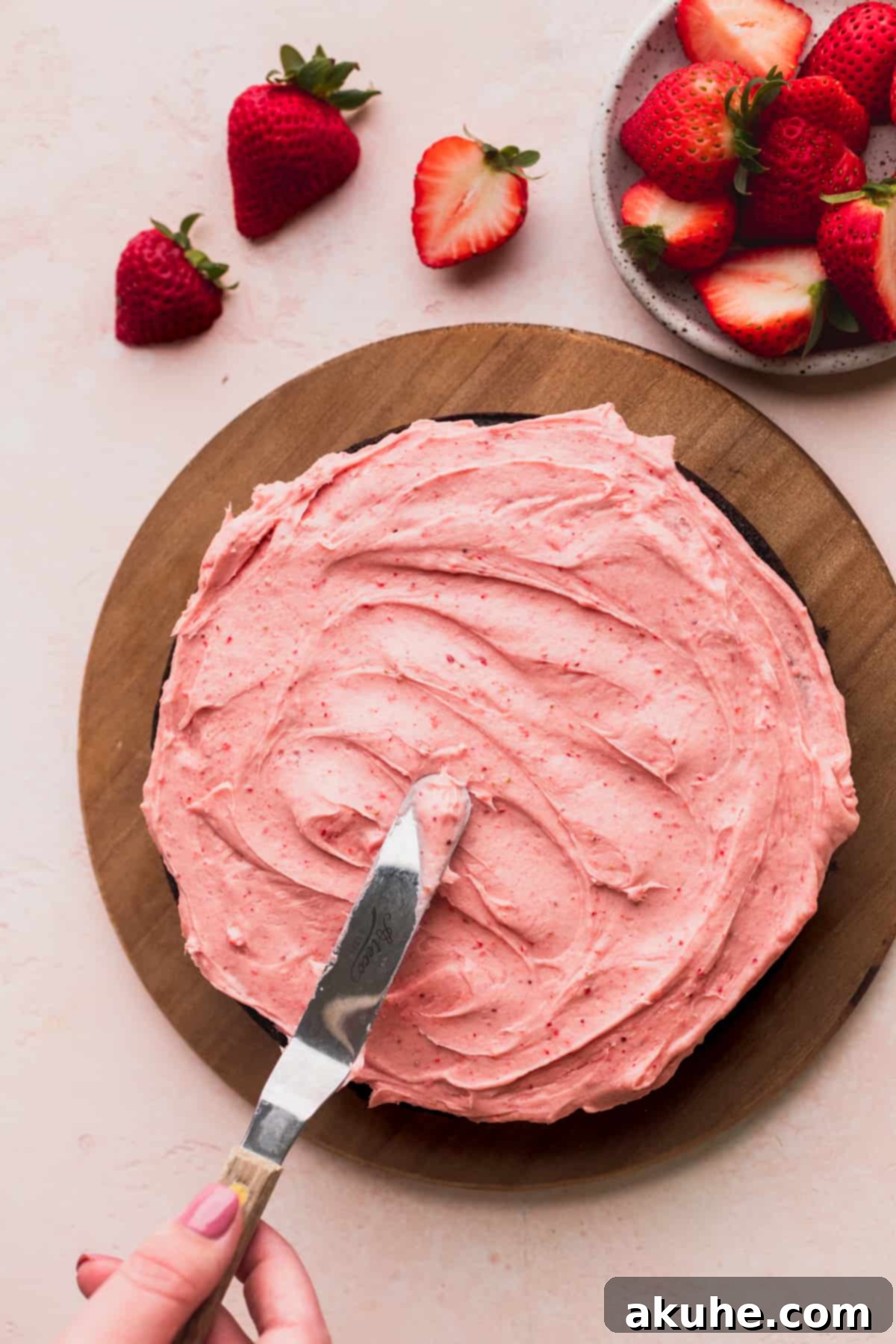 A baker using an offset spatula to evenly spread strawberry frosting on top of a chocolate cake layer.